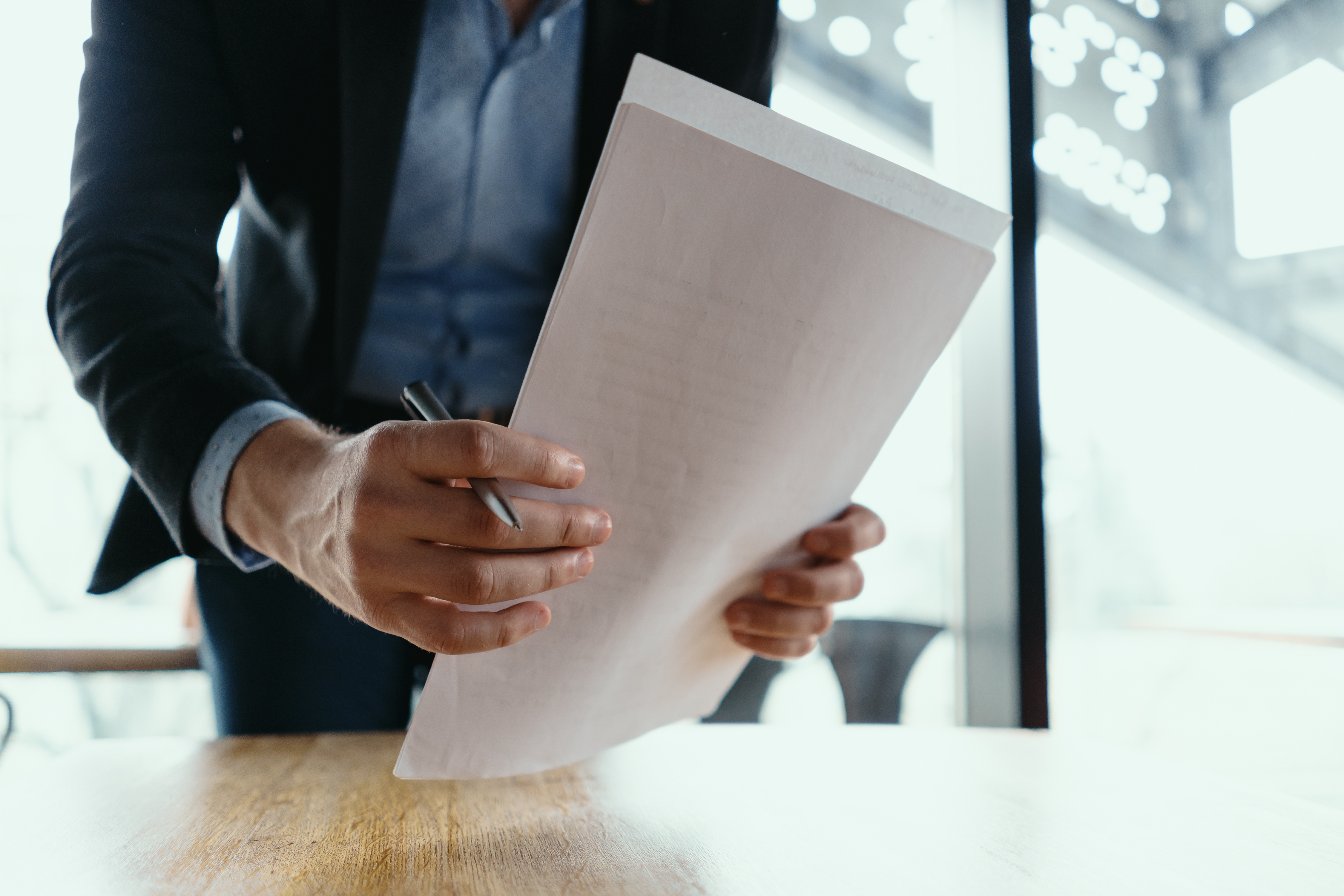 Close up hands signing documents in a modern office with window in background. Pen in hand, papers on the wooden desk, futuristic background.